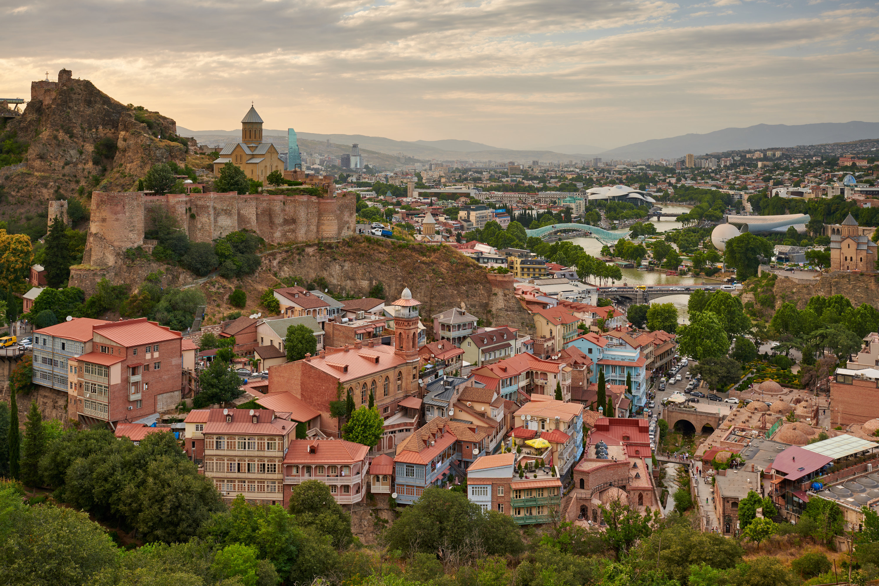 An aerial view of Tbilisi which shows its close and colorful buildings as well as a large river with a bridge in the background