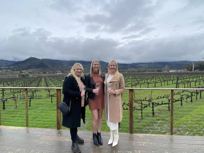 Three women posing for a photo with wine glasses in front of a vineyard