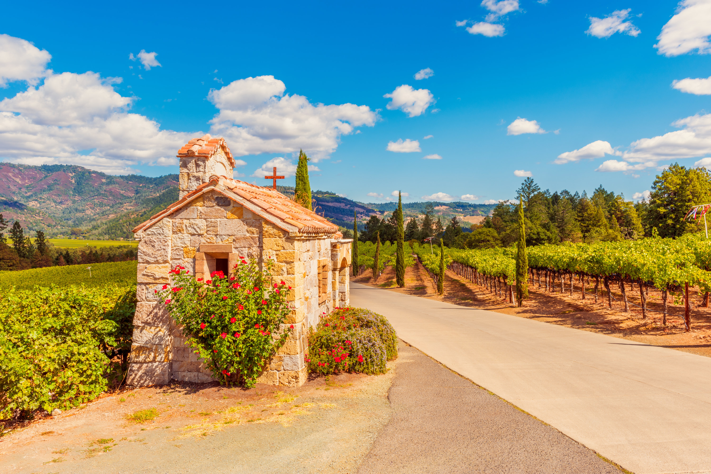 Stone building with cross in front of winery with mountains behind.