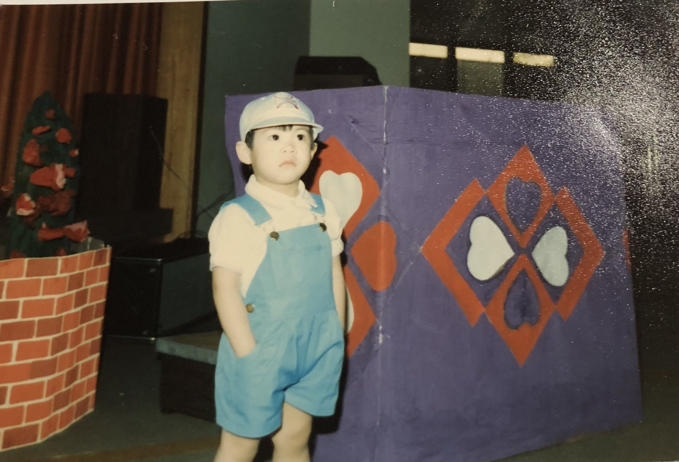 Kim Thai around 5 years old, hands in pocket wearing overalls and a baseball cap.