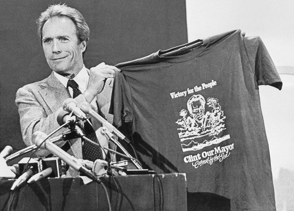 Clint holding up a &quot;Clint Our Mayor&quot; T-shirt at a podium