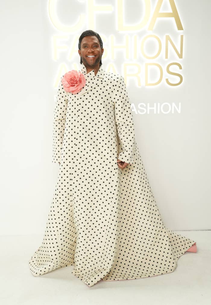 Law Roach attending the CFDA Fashion Awards in November 2022. He wears a long white dress with black polka dots and a large pink peony on his right shoulder. He is smiling.