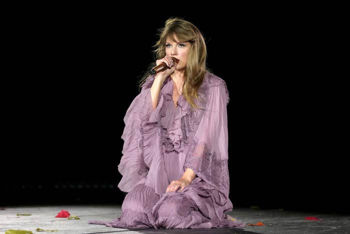 Taylor on her knees as she sings onstage. She&#x27;s wearing a long-sleeve flow-y dress with ruffles around the neck