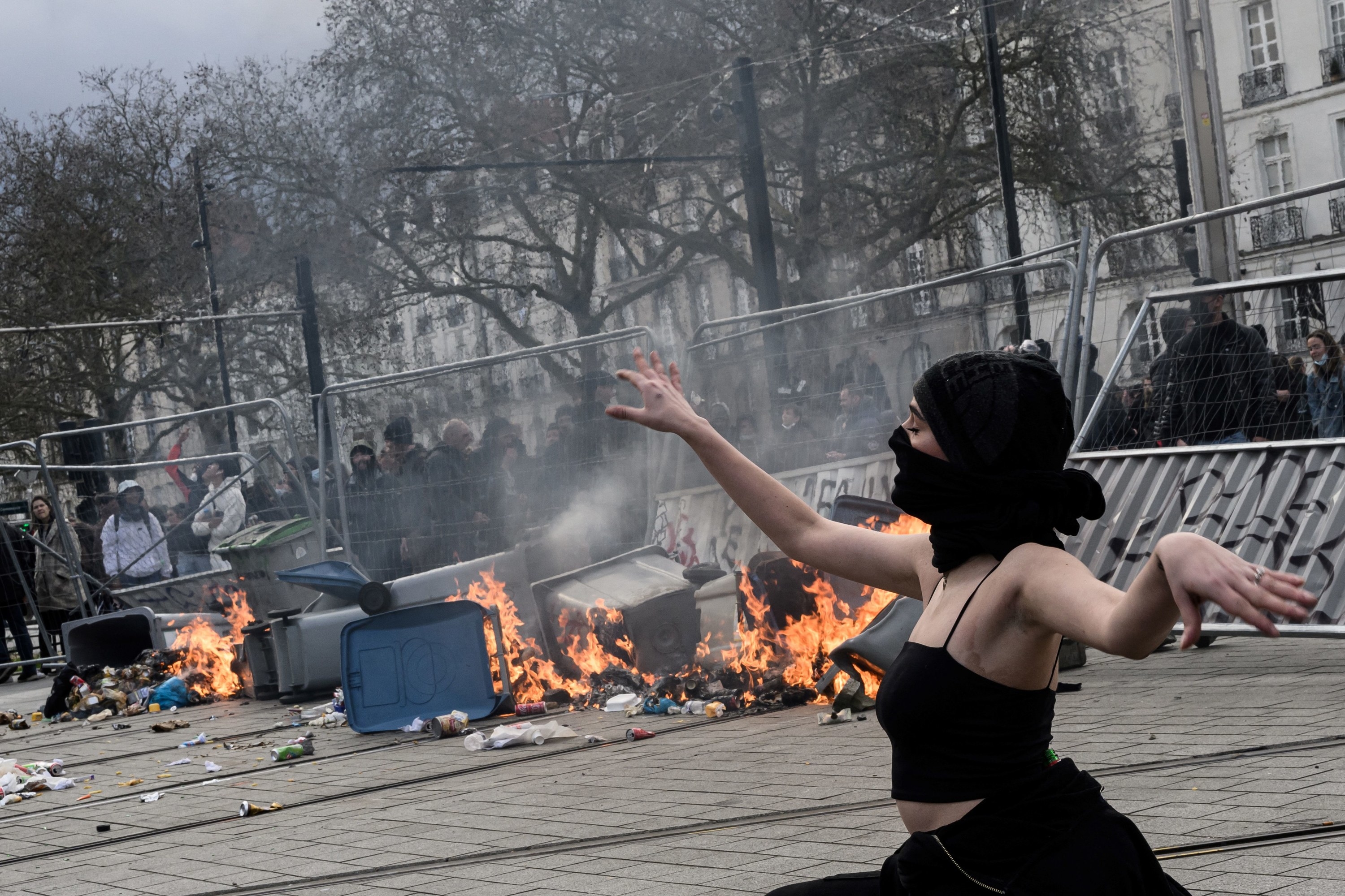 A person wearing a black head covering and mask to obscure their identity poses, as if dancing, in front of a flaming barricade