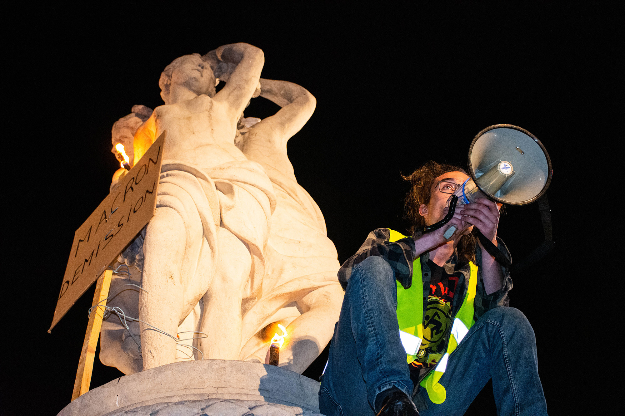 a protester leads the crowd in chants with a bullhorn from the top of a marble statue of three women where a sign says &quot;MACRON DEMISSION&quot; which means &quot;MACRON RESIGN&quot;