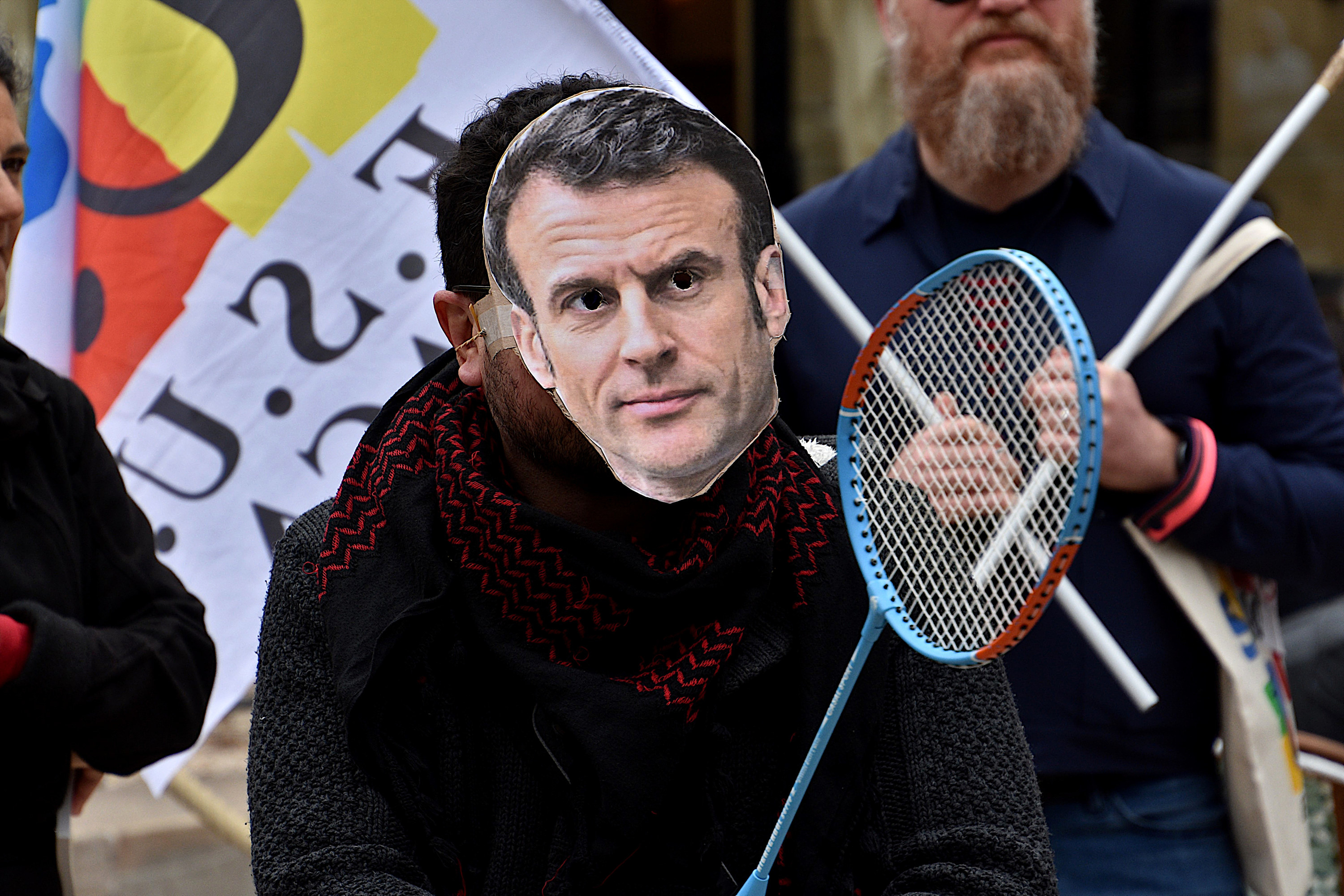 A protester wears a homemade mask with a picture of Macron&#x27;s face and holds a badminton raquet