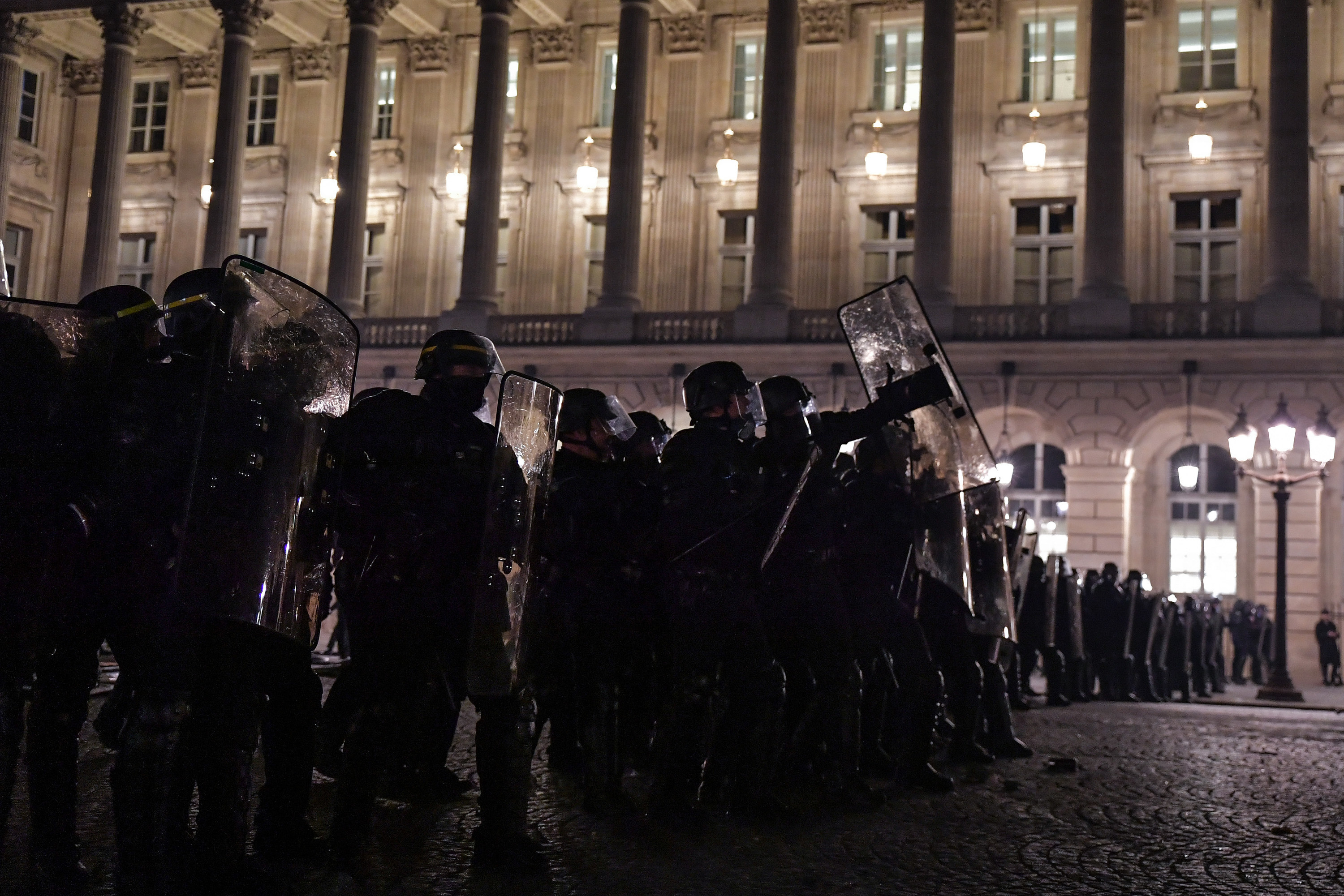 at night, police in riot gear stand in a line holding shields