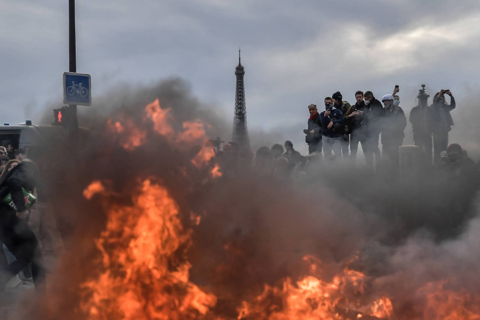 fire burns in the foreground as protesters stand on an elevated surface documenting the scene with the Eiffel Towerin the background