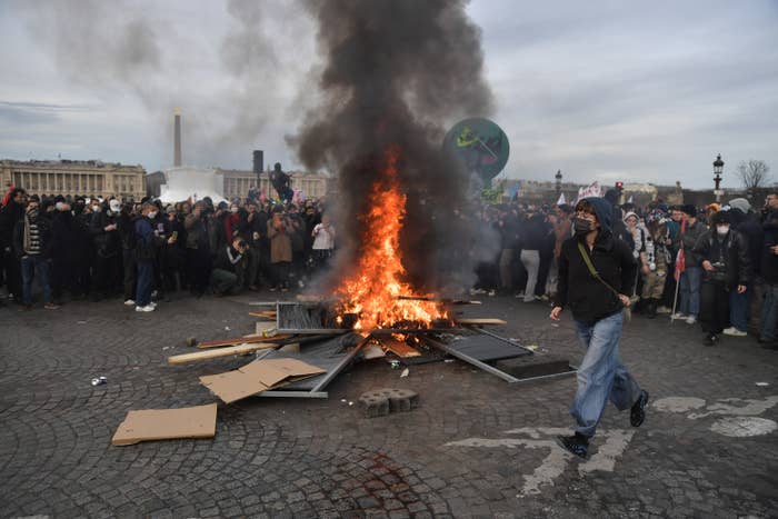 Among a group of protesters, debris is set on fire and a protester in a mask runs by