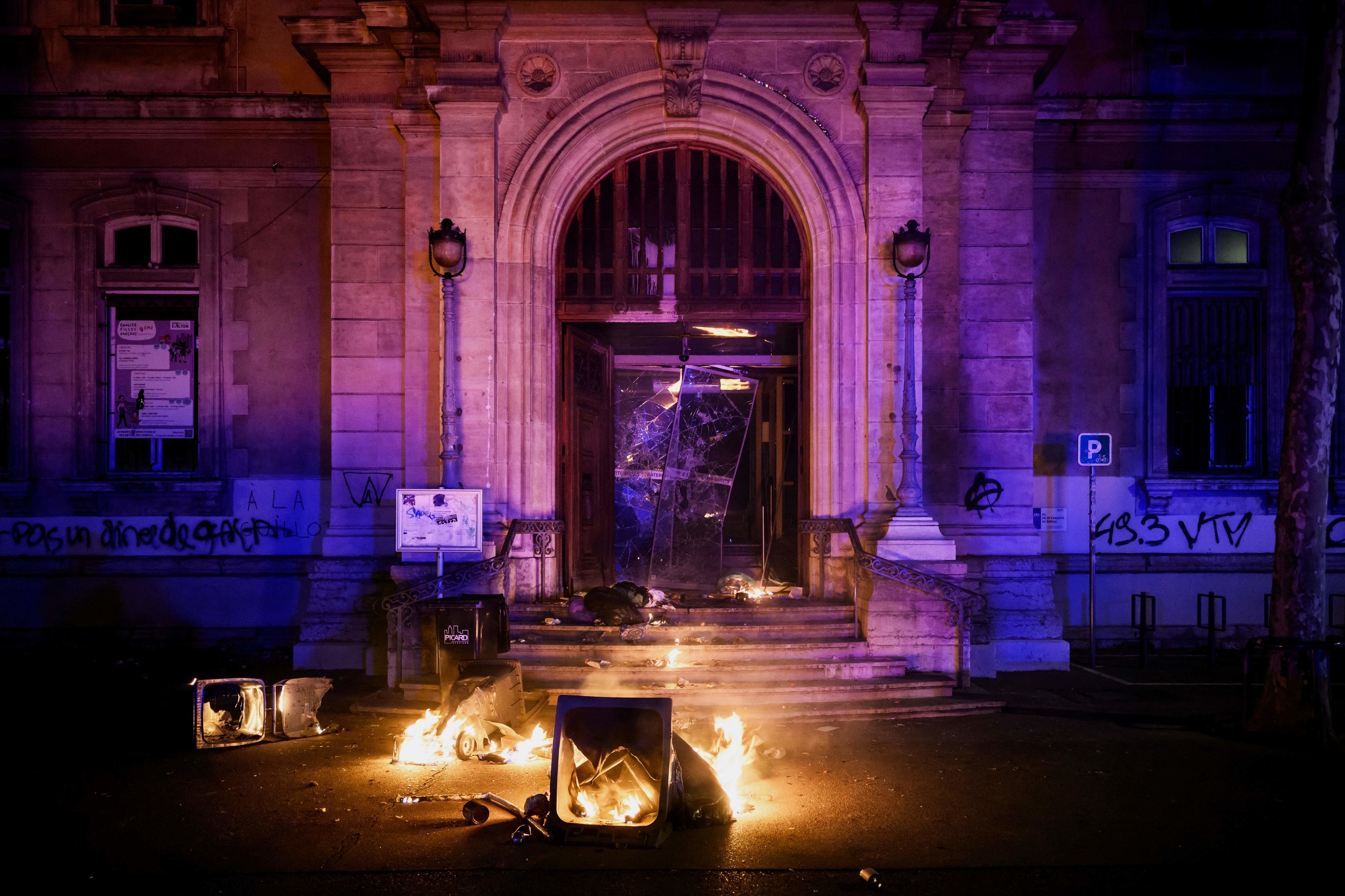 The entrance of a town hall building, marble with decorative filigrees, has been vandalized by spray paint, flaming trash cans, and shattered glass doors