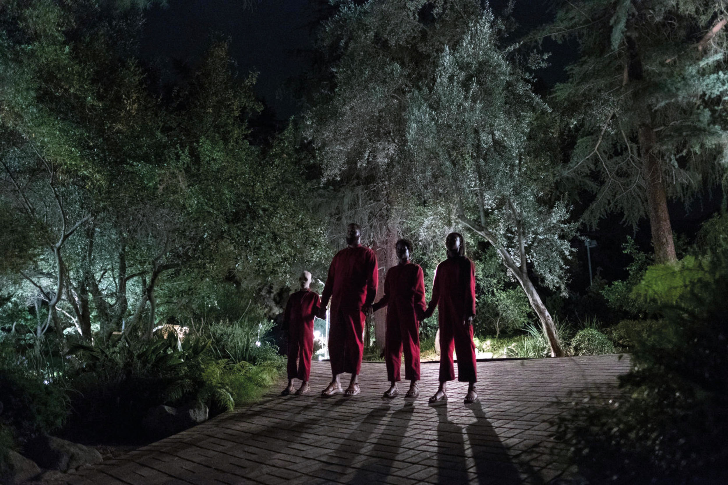 A group of people holding hands in red suits stand ominous in a driveway