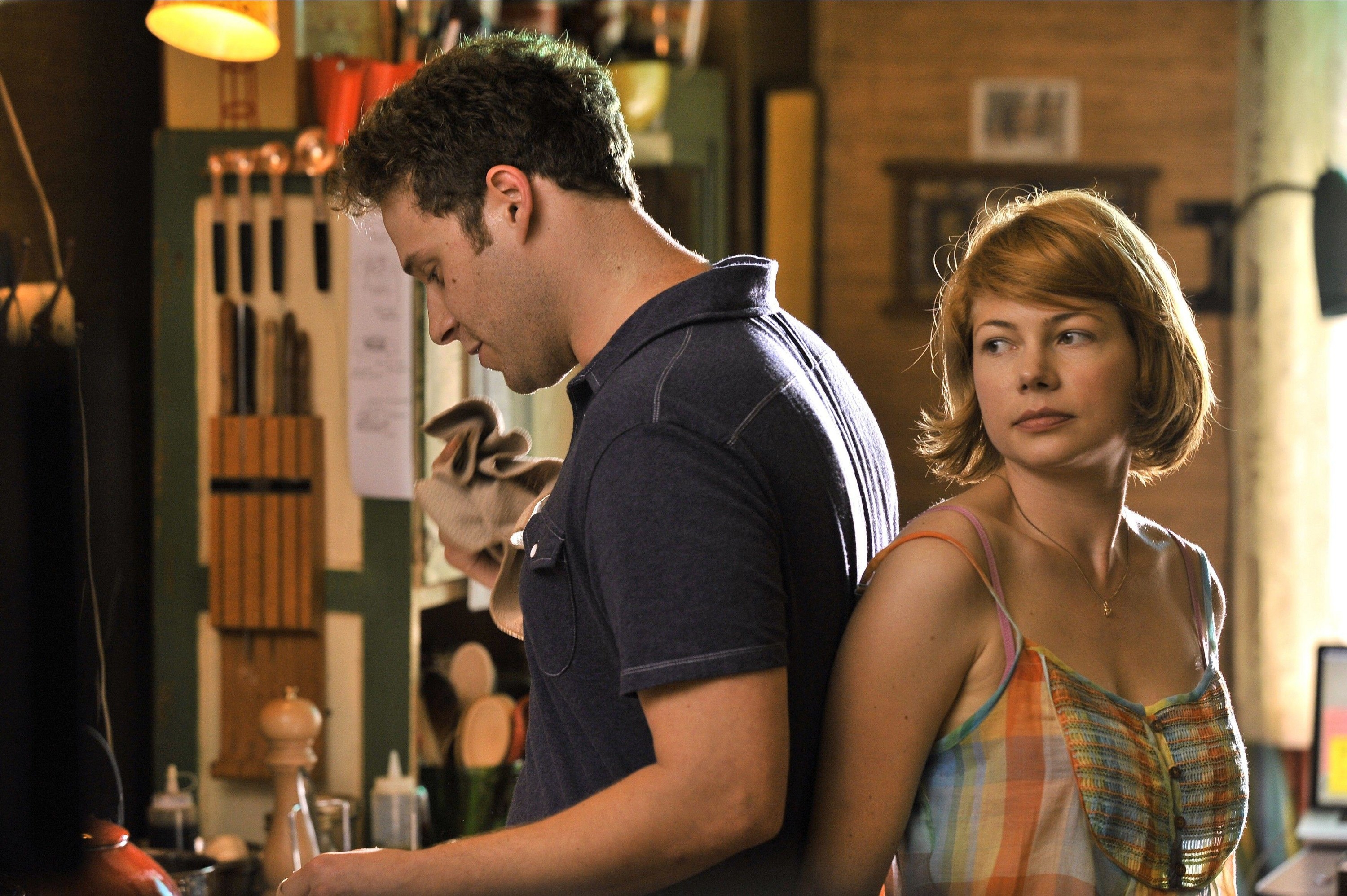 Michelle Williams and Seth Rogen stand back-to-back in a kitchen