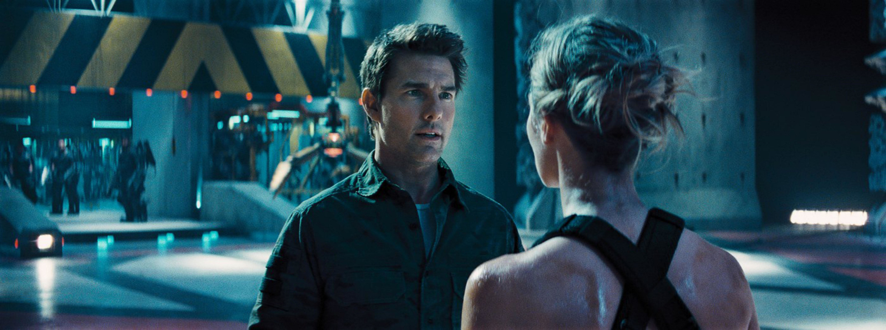 Tom Cruise in a button-up shirt talks to Emily Blunt in a futuristic training facility