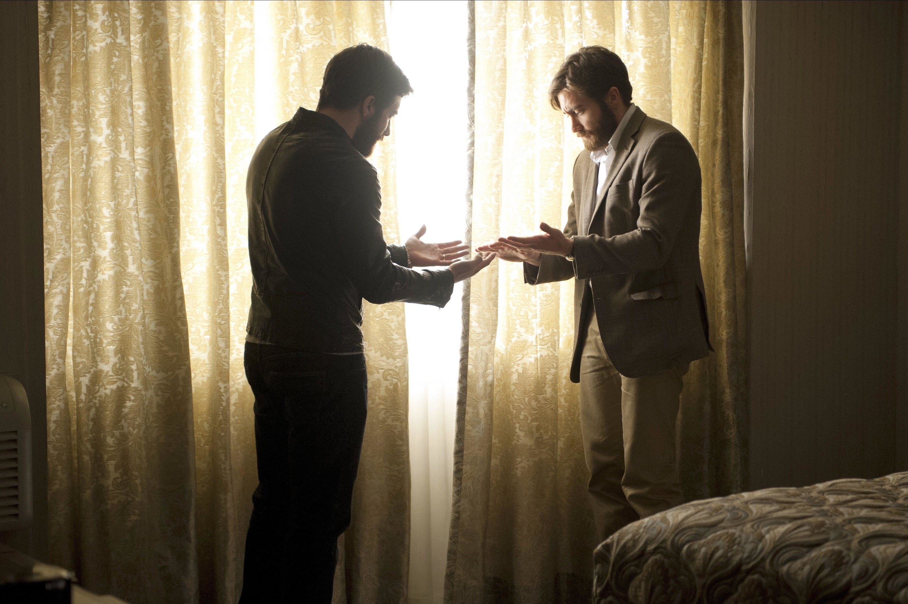 Two Jake Gyllenhaals stand in a dimly lit hotel room