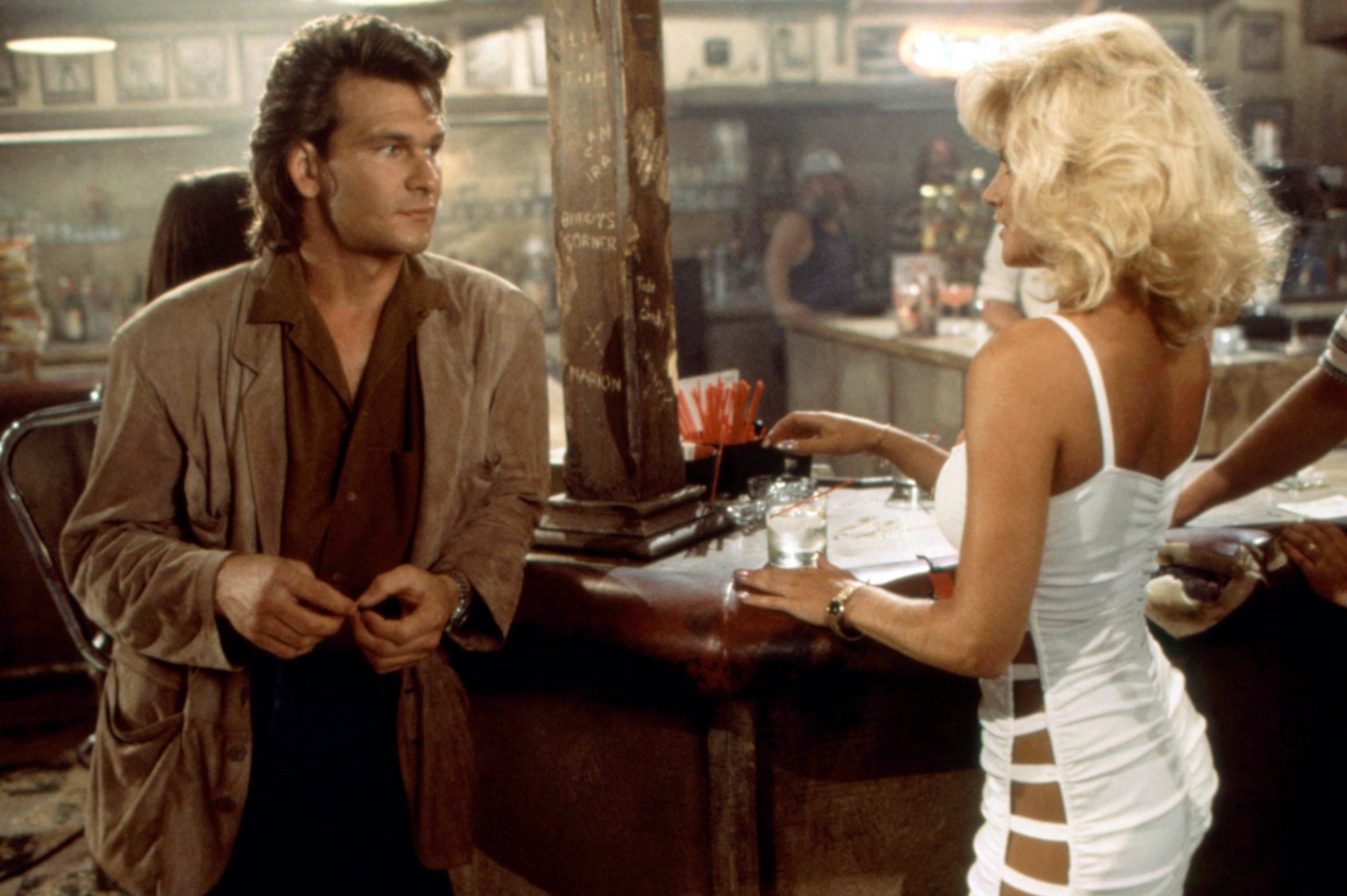 Patrick Swayze leans on a western-themed bar near a blonde woman