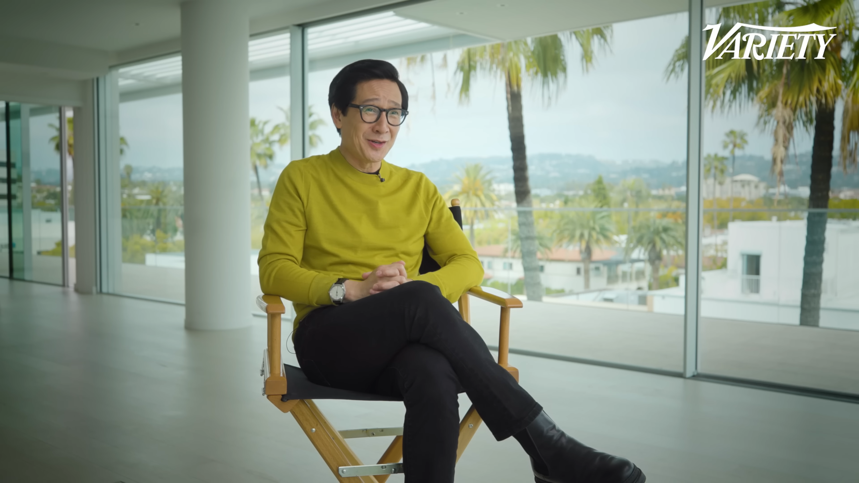 Ke Huy sitting in a director&#x27;s chair during his Variety interview in a room with floor-to-ceiling windows. There are houses and palm trees outside