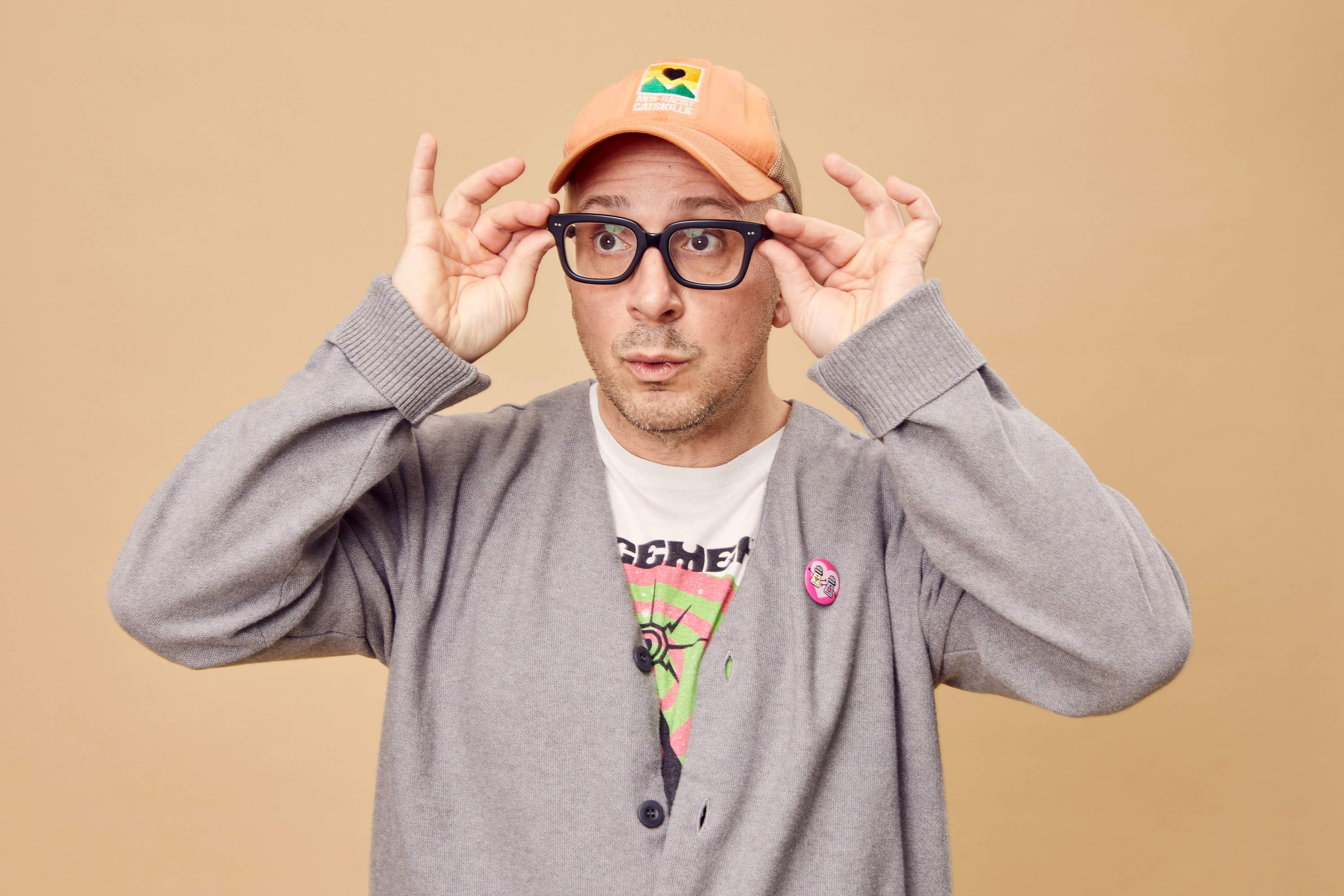 Steve adjusts his eyeglasses as he looks off-camera. He&#x27;s wearing a baseball cap, a band shirt, and a sweater