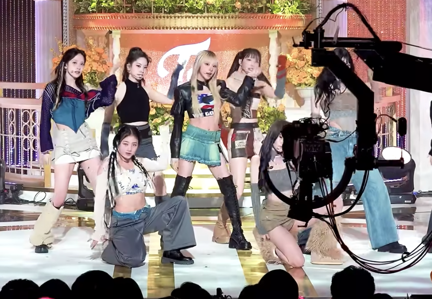 Seven dancers onstage making the same pose, with one blonde dancer in the middle wearing a QAnon T-shirt