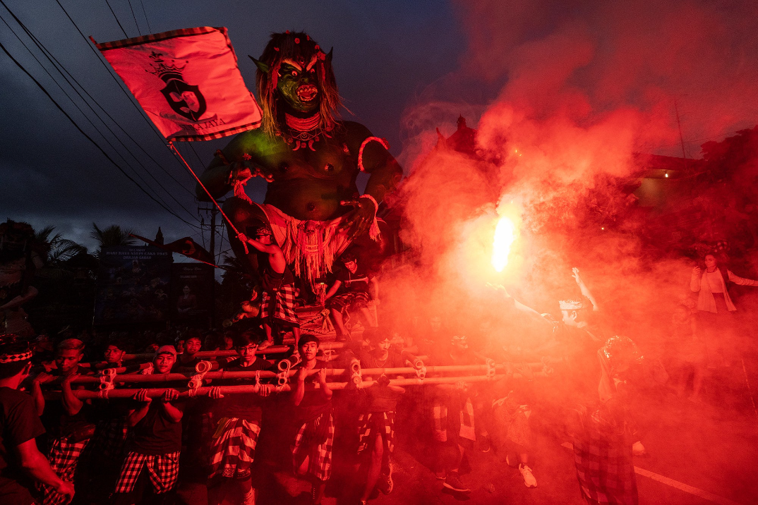 a gigantic green demon effige carrying a flag burns while being held up by a crowd of people holding wooden supports