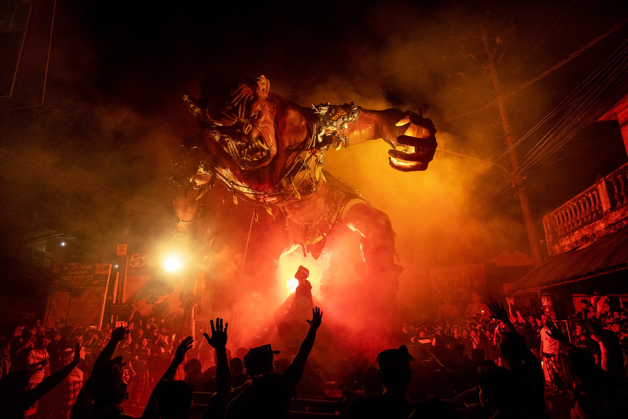a gigantic demon effigy burns and looms over a crowd of onlookers at night