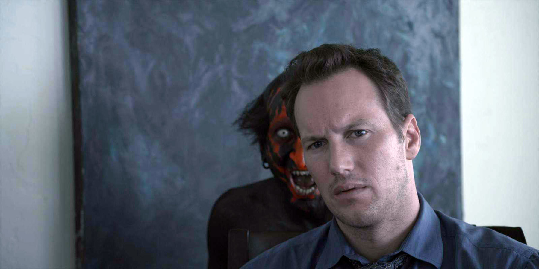 A creature in red face paint lurks behind a seated man