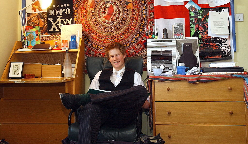 He&#x27;s smiling and sitting in a chair between a small desk and chest of drawers with a psychedelic print above his head