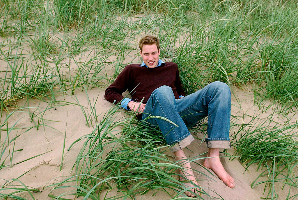 He&#x27;s smiling and leaning back on the grassy sand in jeans and a sweater and barefoot, with his knees bent