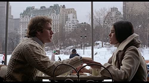 Oliver (Ryan O&#x27;Neal) and Jenny (Ali MacGraw) sit at a table drinking coffee in front of a window with a snowy cityscape outside