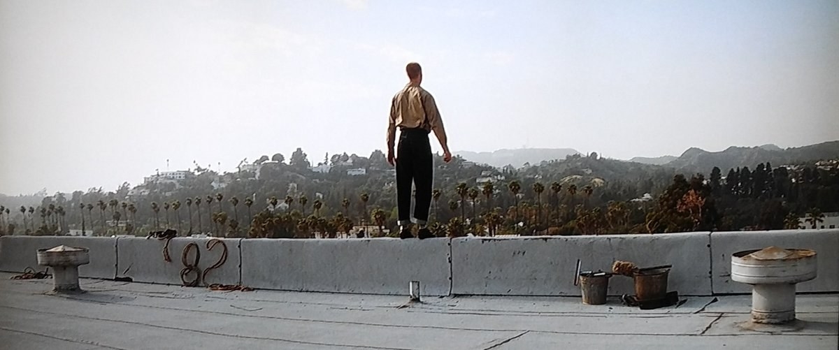 a man stands on a roof overlooking Los Angles, and the number 82 is formed out of copper wire on the ground next to him