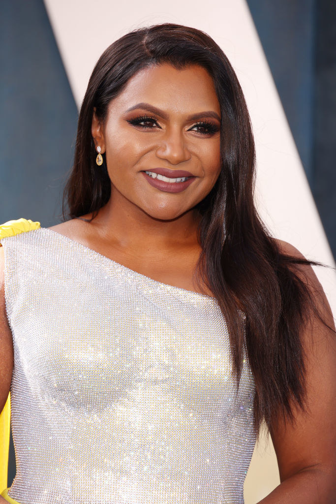 A closeup of Mindy smiling at a red carpet event wearing a one-shouldered sequined dress with a sash at the shoulder