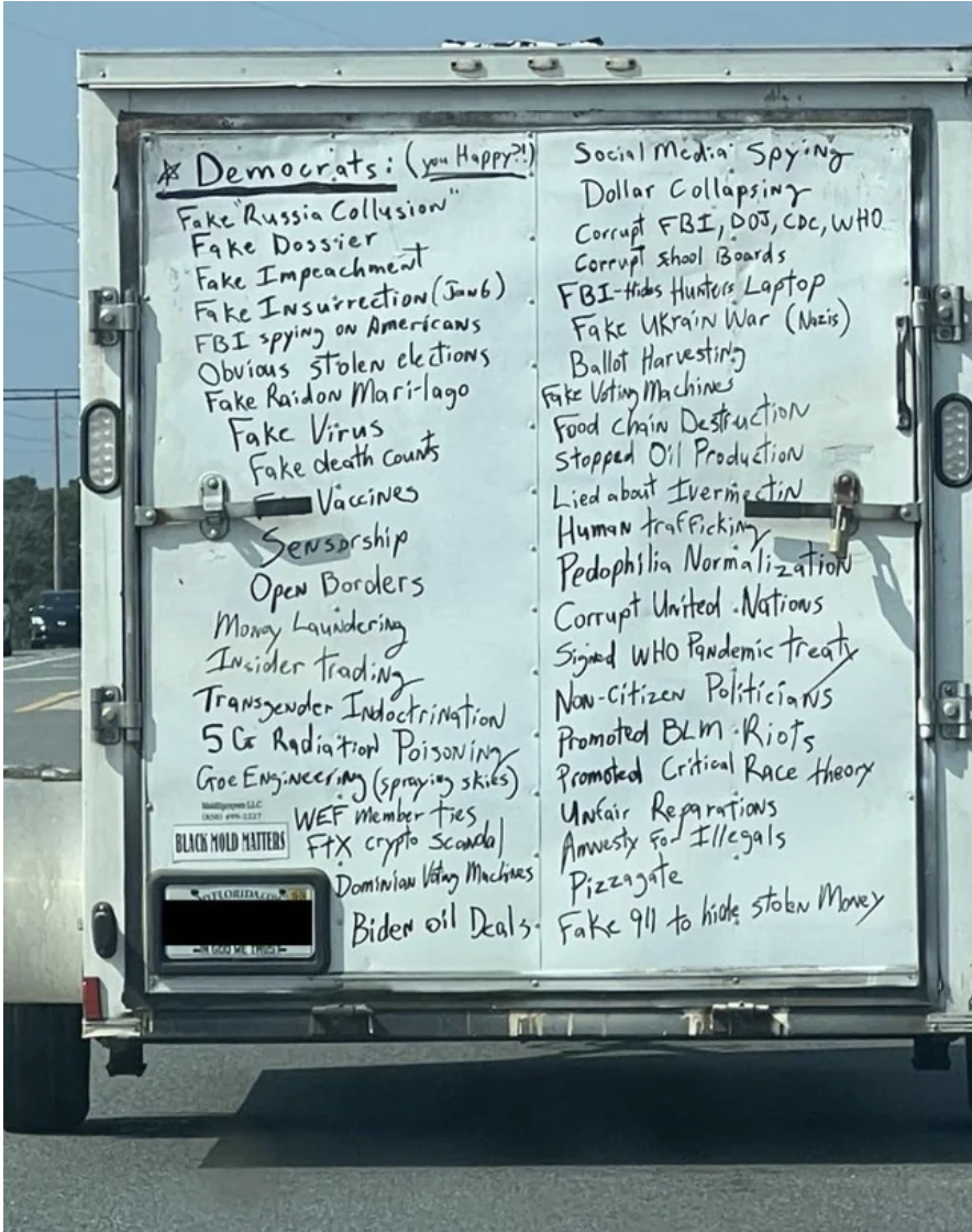 The back of a truck on the highway with a long handwritten list under the heading &quot;Democrats,&quot; including fake dossier, fake virus, fake impeachment, Dominion voting machines, transgender indoctrination, unfair reparations, and pizzagate