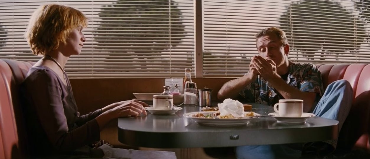 A man and a woman sit at a table in a diner with half-empty plates. The man lights a cigarette.