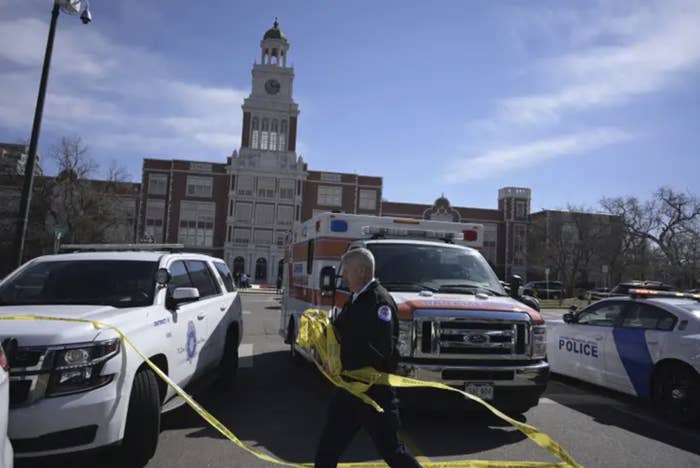 the outside of a high school is flanked by police trucks and a man surrounding the area with caution tape