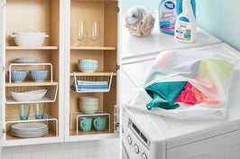 on left, plates and cups stacked on white shelves in cabinet. on right, laundry bag filled with bras on top of washing machine