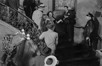 Gloria Swanson as Norma Desmond walks down a grand staircase while surrounded by 1950s-era reporters and police officers