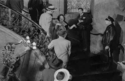 Gloria Swanson as Norma Desmond walks down a grand staircase while surrounded by 1950s-era reporters and police officers