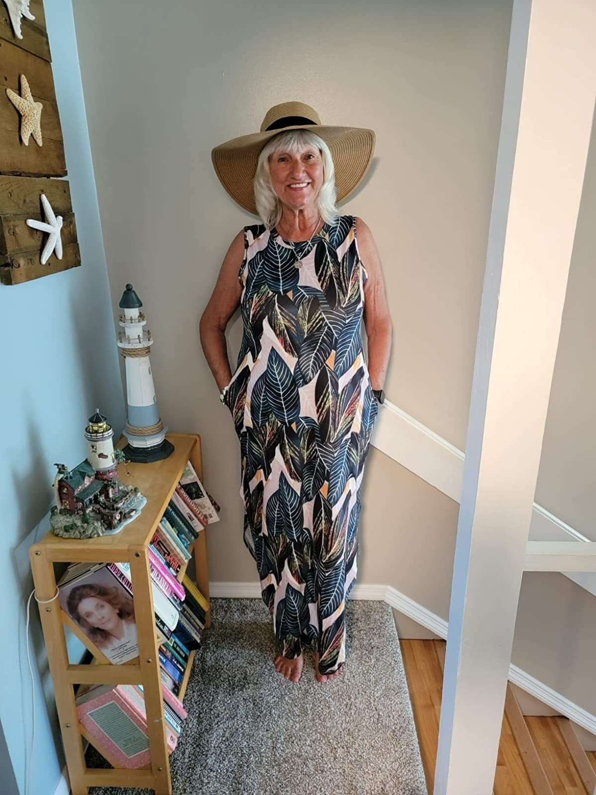 Person smiling, wearing a wide-brimmed hat and a tropical-patterned sleeveless maxi dress, standing near a bookshelf and nautical decor