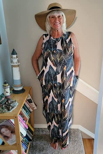 Person smiling, wearing a wide-brimmed hat and a tropical-patterned sleeveless maxi dress, standing near a bookshelf and nautical decor