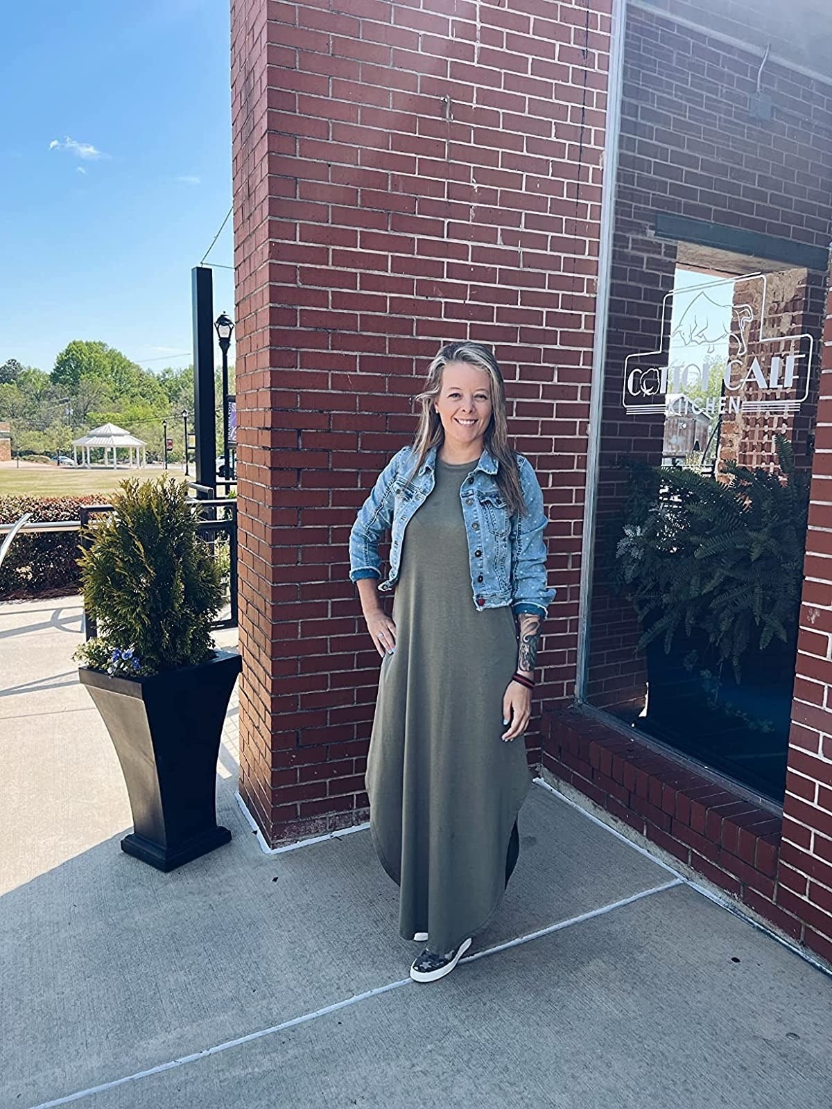 Person in a denim jacket and long dress stands outside a brick building, smiling. Casual attire suitable for everyday wear