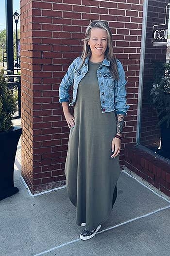 Person in a denim jacket and long dress stands outside a brick building, smiling. Casual attire suitable for everyday wear