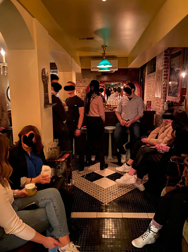 People (with their faces blacked out) standing or sitting around in a small room with a bar