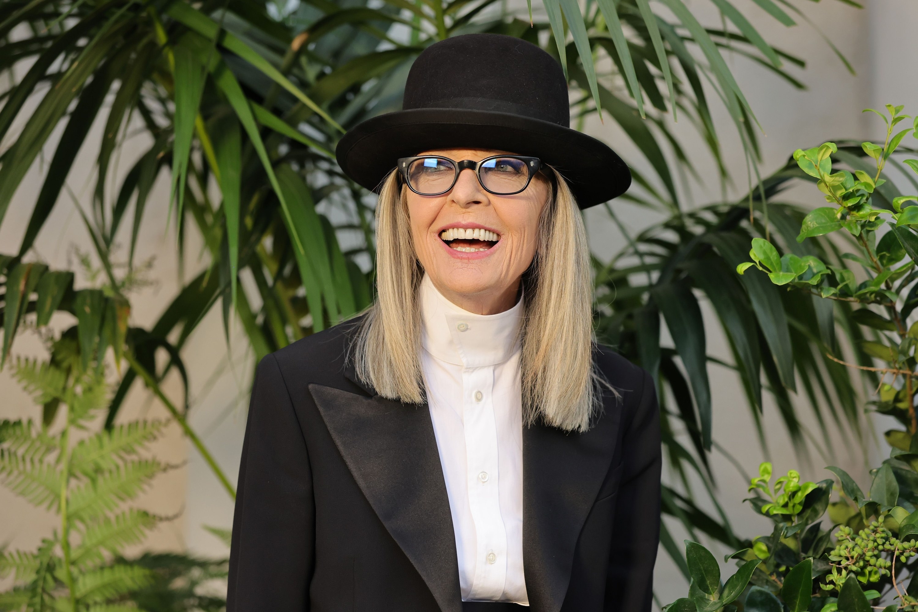 A closeup of Diane smiling. Diane is wearing her usual pantsuit and hat combo