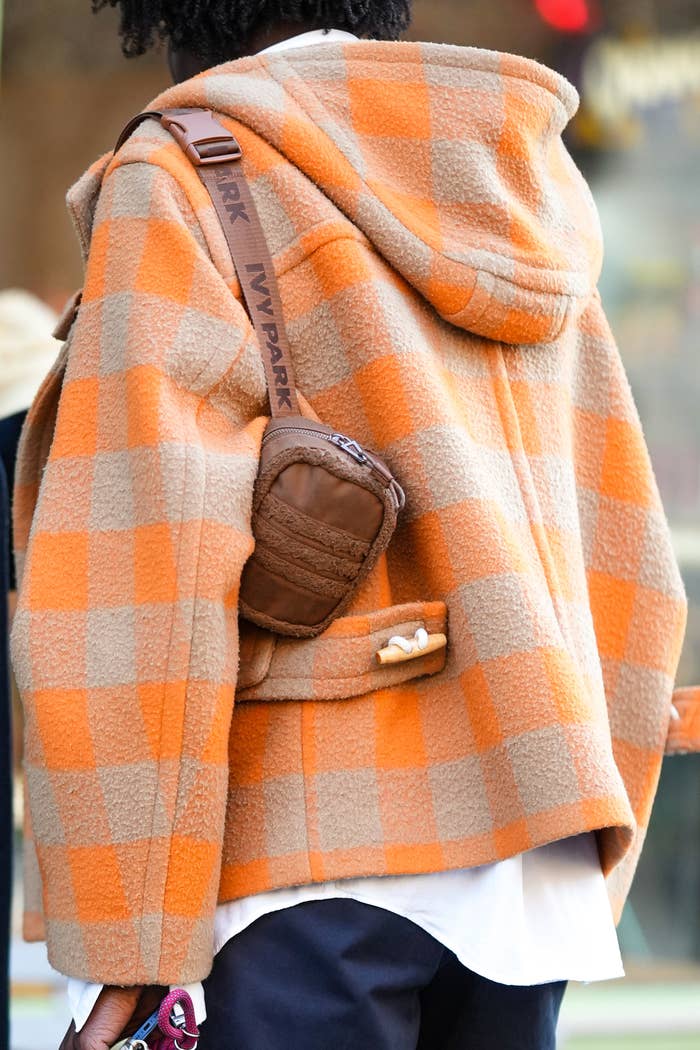 Someone with their back to us wears a white shirt, a beige and orange checkered print pattern wool hoodie coat, black suit pants, a brown nylon and fluffy shoulder bag