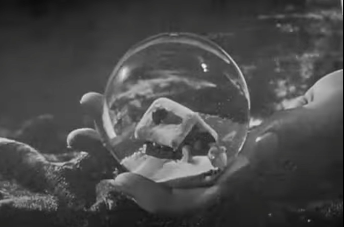 a black and white image of a hand holding a snow globe that contains a small snowy cabin
