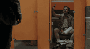 Ryan Gosling, holding a gun, drops a cigarette in his pants while on the toilet