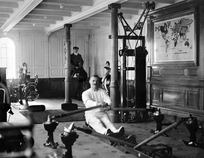 Black-and-white photo of a man in a rowing machine in a large room with a map of the world on the wall
