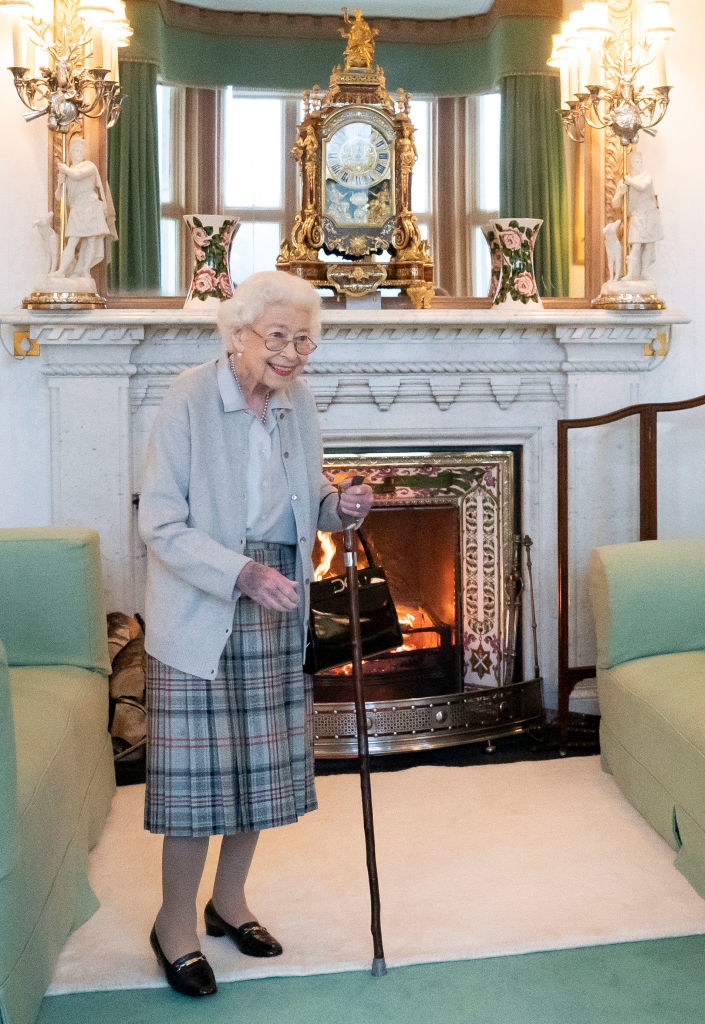 The Queen standing and smiling in front of a fireplace with a cane