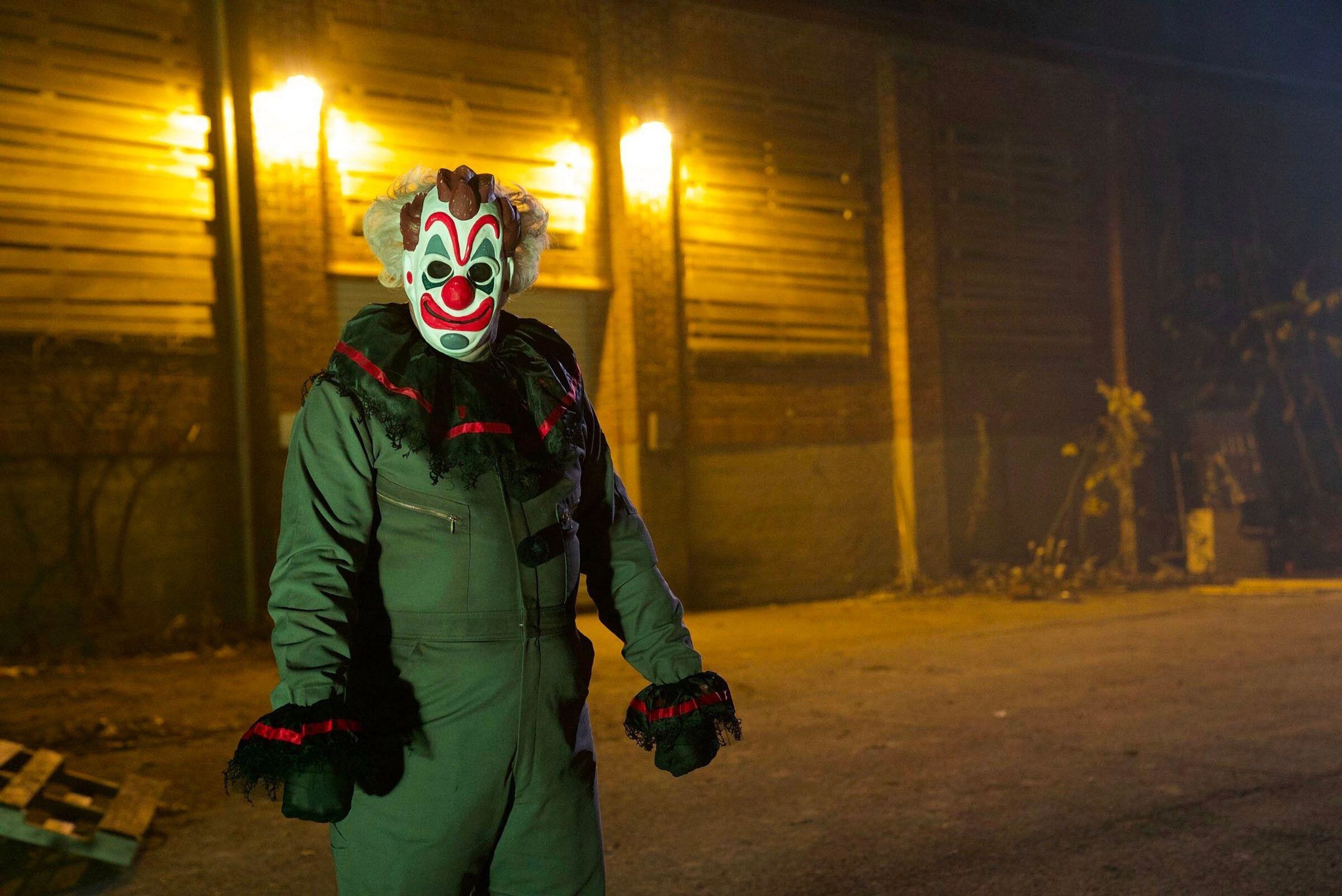 A menacing man in a clown mask stands outside a deserted warehouse