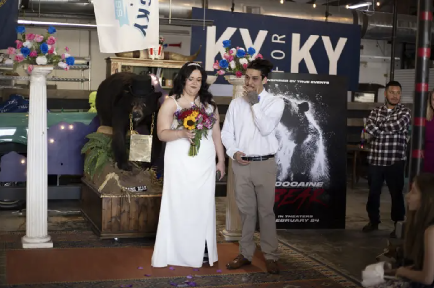 a man and woman in wedding formalwear stand in front of a stuffed black bear and a poster for the cocaine bear movie inside a mall
