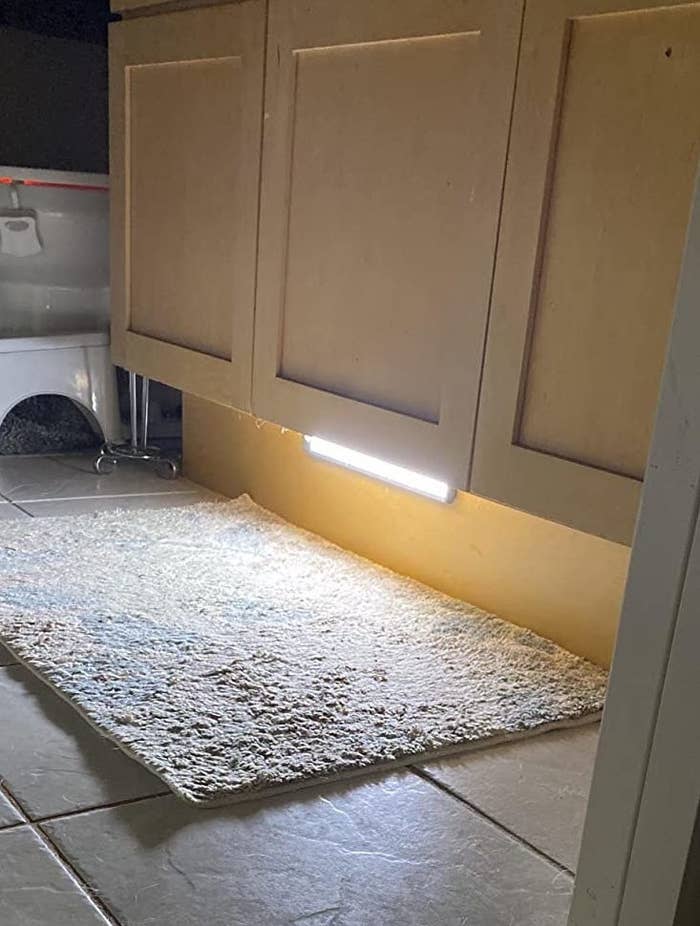 Under-cabinet LED light illuminating a kitchen mat on a tiled floor, with partial view of cabinets and a container